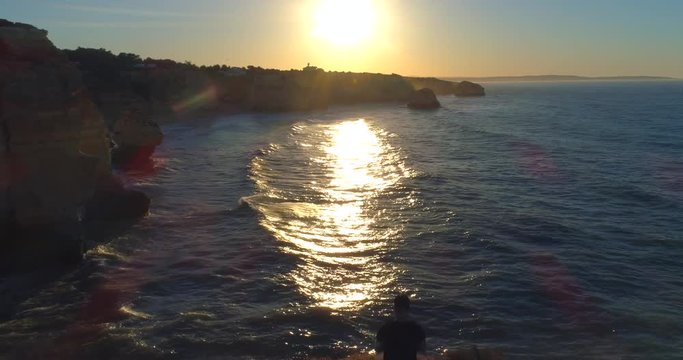 Aerial, reverse, drone shot,  silhouette, over a man standing on a cliff, at the sea, Heart rocks arch, at the Marinha beach, at sunrise, on a sunny, morning dawn, in Carvoeiro, Algarve, Portugal