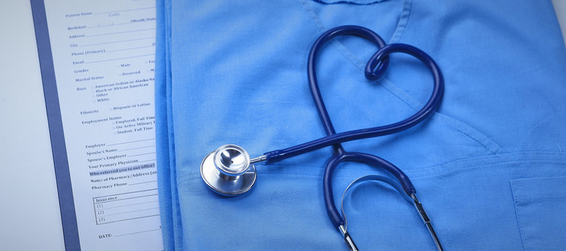 A medical stethoscope is intertwined in the shape of a heart and lies on a medical history and a blue uniform. Closeup of a doctor's uniform.