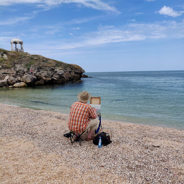 artist paints a landscape from nature on the shore of the azure sea. artist in a straw hat with malbert draws on canvas