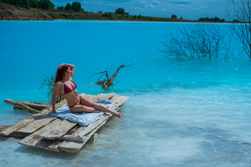A beautiful red-haired woman in a seductive crimson bikini sits in front of an unusual turquoise lake. A miracle of nature, bright blue clear water. Ash dump, poisoned pond.