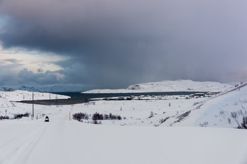 Snow covered winter road lead to a small fishing village on the sea in the far North, above the Arctic Circle in Russia's Kola Peninsula. Small snowmobile on the road.