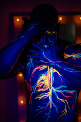 Fototapeta na wymiar UV patterns body art of the circulatory system on a man's body. On the torso of a muscular athlete, veins and arteries are drawn with fluorescent dyes. Bodybuilder standing by the mirror with lamps.