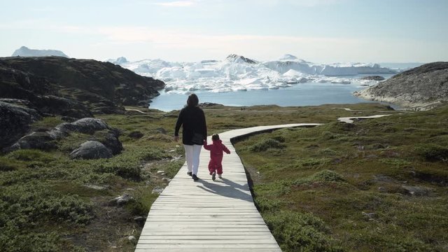 POV: Woman and Toddler Walk on Wooden Walkway in Lush Field Towards Ice Fjord - Disko Bay, Greenland