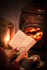 Woman/girl sitting in front of a cozy fireplace during winter under a blanket  reading a book drinking coffee/hot chocolate.