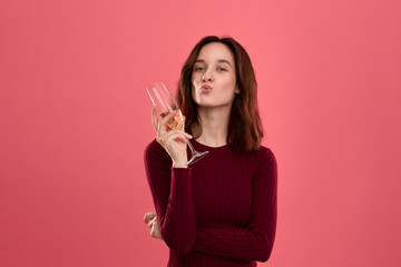 Excited pretty brunette girl holding wineglass of sparkling champagne standing isolated on a dark pink background and smiling at the camera. Model sending air kisses.