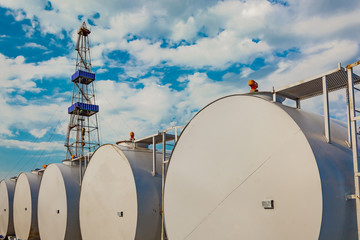 In the foreground are gray cylindrical tanks for storing oil and fuel reserves. Located horizontally in a row. In the background is a rig for drilling oil wells. Blue cloudy sky.