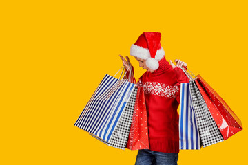 Cute boy in red sweater holding shopping bags with Christmas gifts and surprised looking in it on yellow background