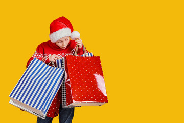 Cute boy in red sweater holding shopping bags with Christmas gifts and surprised looking in it on yellow background