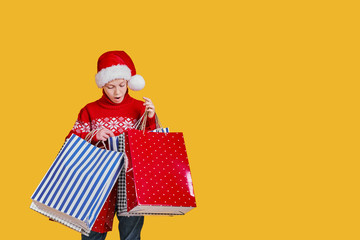 Cute boy in red sweater holding shopping bags with Christmas gifts and surprised looking in it on yellow background