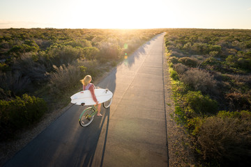 Beautiful blonde surfer girl on her way to the beach on her bicycle with her surfboard.