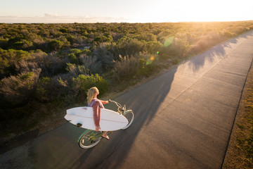 Beautiful blonde surfer girl on her way to the beach on her bicycle with her surfboard.