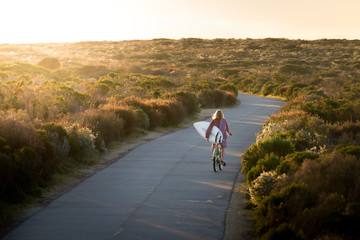 Beautiful blonde surfer girl on her way to the beach on her bicycle with her surfboard.