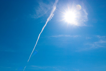 Trail from the plane against the blue sky with cirrus clouds and bright sun.