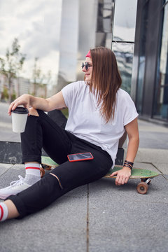 Image of young brunette woman in sunglasses with glass in her hands sitting on skateboard on background of modern buildings in city