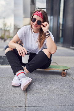 Image of young brunette woman in sunglasses looking at camera with glass in her hands sitting on skateboard on background of modern buildings in city
