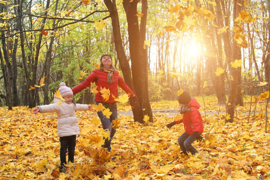 Happy Family Walking In Sunny Park And Throws Orange Maple Leaves. Mother With Kids Enjoying Autumn Weather Outdoors