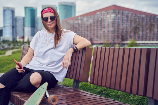 Photo of woman skateboarder with phones in her hands sitting on wooden bench against backdrop of modern buildings