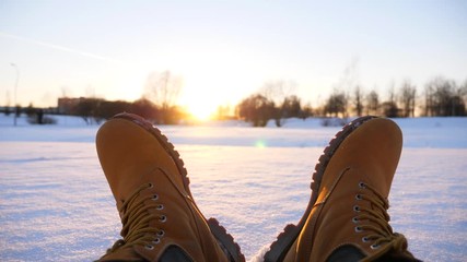 Warm leather shoes on snow, sun light shine low at evening time. POV shot of male relaxing sitting on frosty snow. Man looking on footwear against sunset