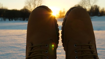 Toes of winter boots against setting sun, man stretch out legs on snow, close shot of warm leather winter shoes. Cold season at European country, blurred city park seen on background, setting sun beam