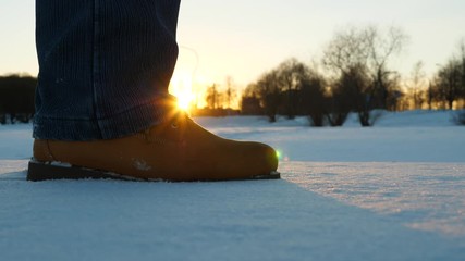 Man step at snow and stand, rise up on tiptoe. Close up shot of warm winter boots against setting sun and blurred city park on background