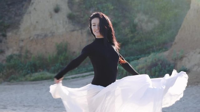 beautiful smiling dark haired girl in bodysuit and long skirt dances on empty sea coast at bright sunlight slow motion