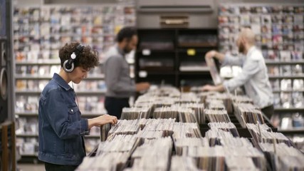 Medium shot of multiethnic customers browsing large assortment of cds and vinyls in record store, chatting and showing each other discs, while curly Caucasian woman in headphones is swaying to music - Powered by Adobe