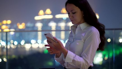 Business woman using smartphone, stay at rooftop terrace, night time, half length portrait. Blurred city lights on background, lady focused on mobile device screen, running application