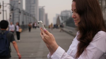 Woman using phone, type text message, blurred pedestrian bridge on background. Brunette lady chatting in instant messenger, stay at footbridge. Citizens silhouetted seen on background out of focus