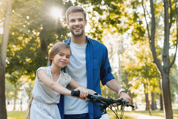 Cute smiling girl learning to ride a bicycle with her father outdoors with look in camera