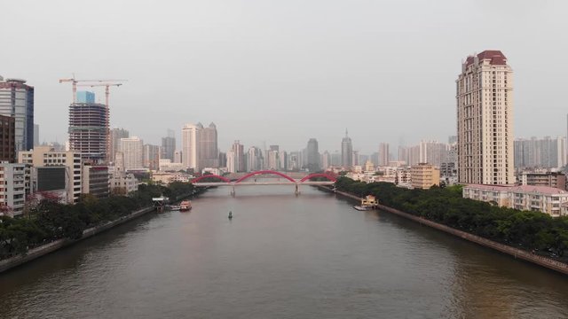 Green banks of Pearl river, red Jiefang bridge span across water, quick aerial shot of Guangzhou city. Tall building on right side, tower under construction on left bank.