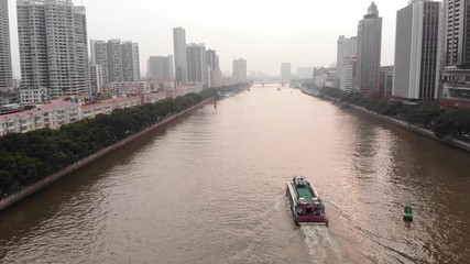 One ferry sail at Pearl River, aerial follow camera, tall buildings on banks, green trees grow along water. Typical light mist in air at evening time. Many tall and low-rise buildings around river