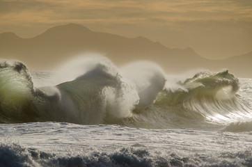 Large wedging waves hitting the coastline under soft sunset light.