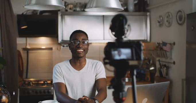 Happy young smart black blogger man filming new vlog video with professional camera in kitchen at home slow motion.