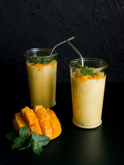 mango smoothie / mango lassi in vintage glass cups on a dark surface with metal straws. with mint leaves and a slice of ripe and juicy mango