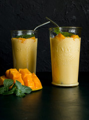 mango smoothie / mango lassi in vintage glass cups on a dark surface with metal straws. with mint leaves and a slice of ripe and juicy mango