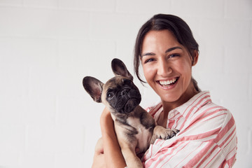 Studio Portrait Of Smiling Young Woman Holding Affectionate Pet French Bulldog Puppy