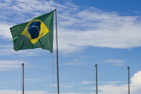National flag of Brazil, swaying in the wind, next to three other empty flagpoles, with a slightly cloudy blue sky background.