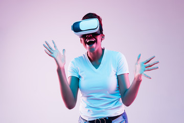 Portrait of young african-american woman's playing in VR-glasses in neon light on gradient background. Concept of human emotions, facial expression, modern gadgets and technologies. Look astonished.