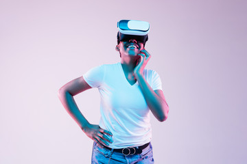 Portrait of young african-american woman's playing in VR-glasses in neon light on gradient background. Concept of human emotions, facial expression, modern gadgets and technologies. Look happy, smiles