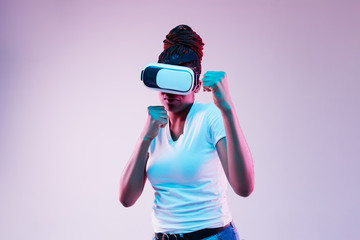 Portrait of young african-american woman's playing in VR-glasses in neon light on gradient background. Concept of human emotions, facial expression, modern gadgets and technologies. Fighting.