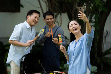 Selfie time! Asian senior group friends making selfie and smiling while having fun grilling meat, cooking enjoying barbecue party in nature. People, Retirement, happy, reunion or Aged Society concept
