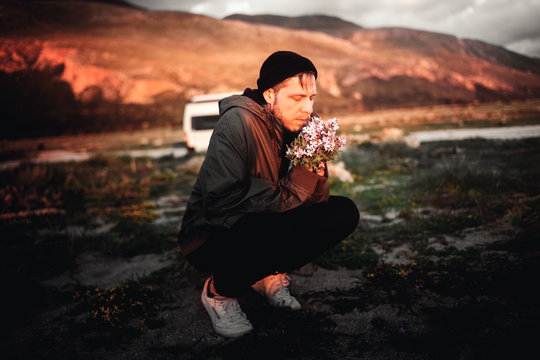 Mann kniet am Strand mit Blumen in der Hand, im Hintergrund ein Campervan und Berge