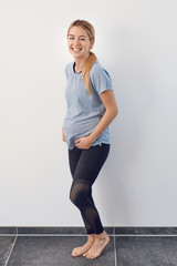 Cute charismatic young pregnant woman standing in front of a white wall with bare feet and hands cradling her belly grinning happily at the camera