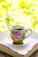 Hot morning coffee in small floral design cup on shabby chic vintage white tray on window sill in front of summer window