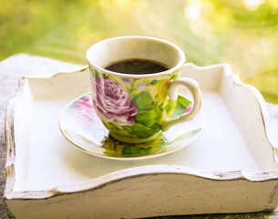 Hot morning coffee in small floral design cup on shabby chic vintage white tray on window sill in front of summer window