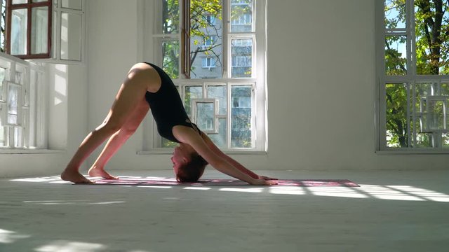 Young red-haired skills healthy caucasian woman practicing yoga on the mat in the white large room. Young woman in sports clothing exercising on fitness indoors.
