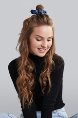 Cropped shot of a ginger girl with wavy hair, sitting on a floor. She wearing black turtleneck and blue jeans. Girl has blue hairpin on her head. Her hair is pulling in high side bun. 