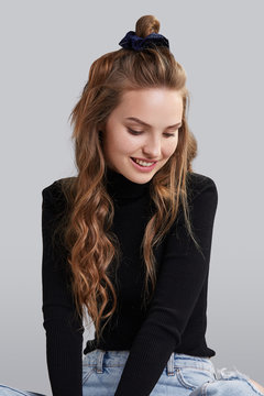 Cropped shot of a ginger girl with wavy hair, sitting on a floor. She wearing black turtleneck and blue jeans. Girl has dark blue hairpin on her head. Her hair is pulling in high side bun. 