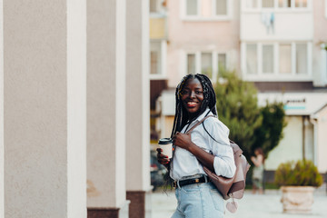 Portrait of a smiling young African American girl with pigtails with coffee walking in the street on a sunny day. Outdoor photo.