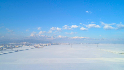 Fototapeta na wymiar AERIAL: Flying above white snow fields towards rocky mountains on winter morning